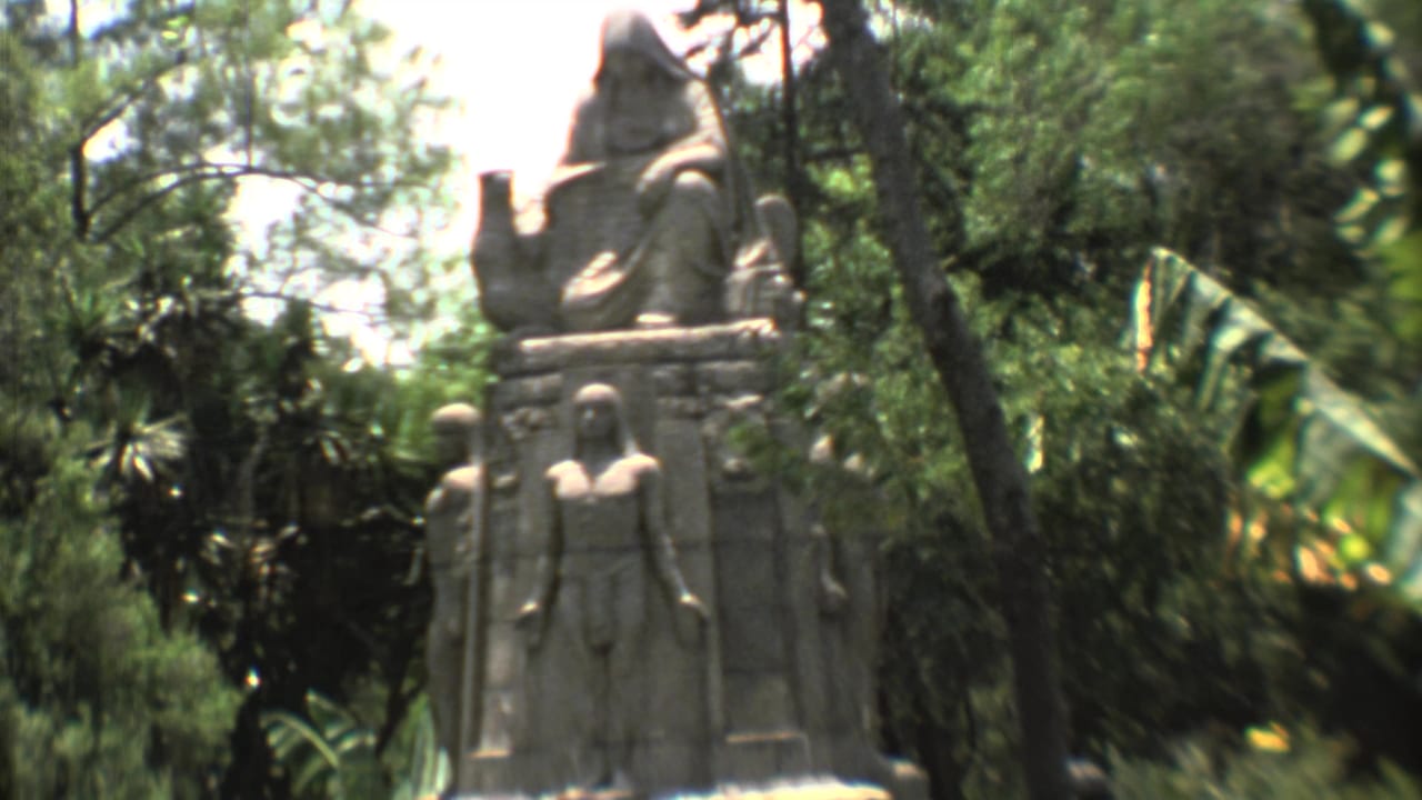 Carved stone monument with seated figure amid dense trees and tropical foliage.