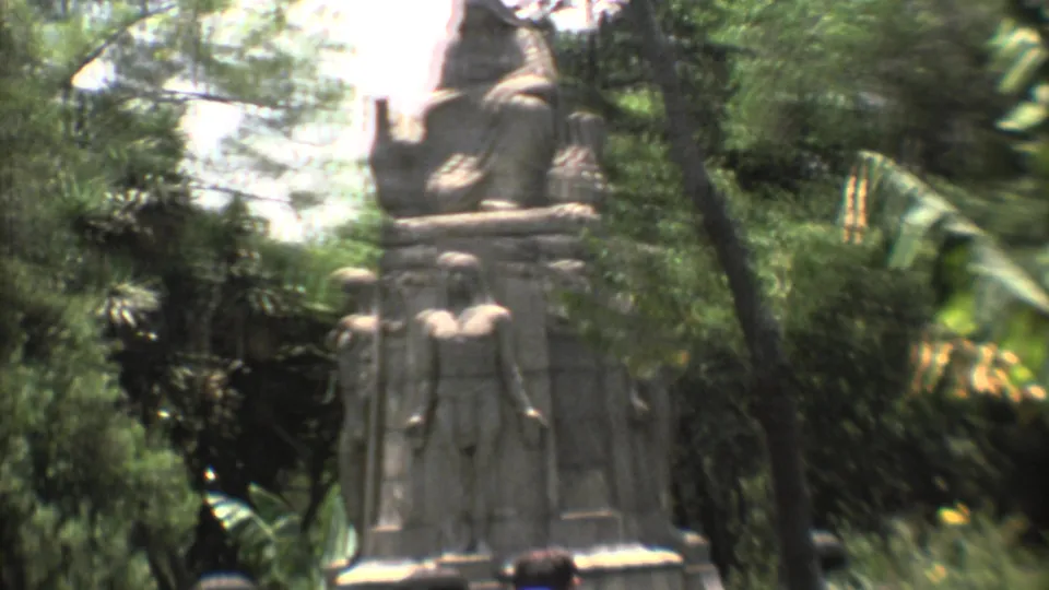 1973 Mexico City frame with carved stone figures seen through leafy garden foliage in soft 8mm color.