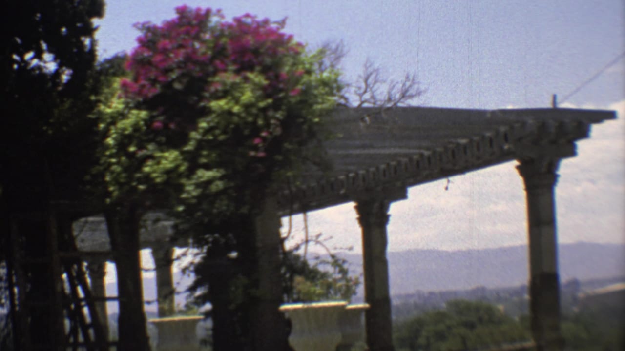 1973 Mexico City home-movie frame showing a sunlit veranda, classical columns, bougainvillea, and a distant hillside view.