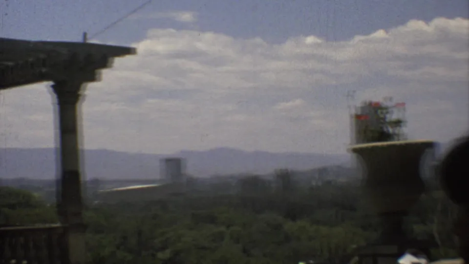 Mexico City overlook from a classical porch, with a column, urn planter, green canopy, high-rise forms, and distant mountains.