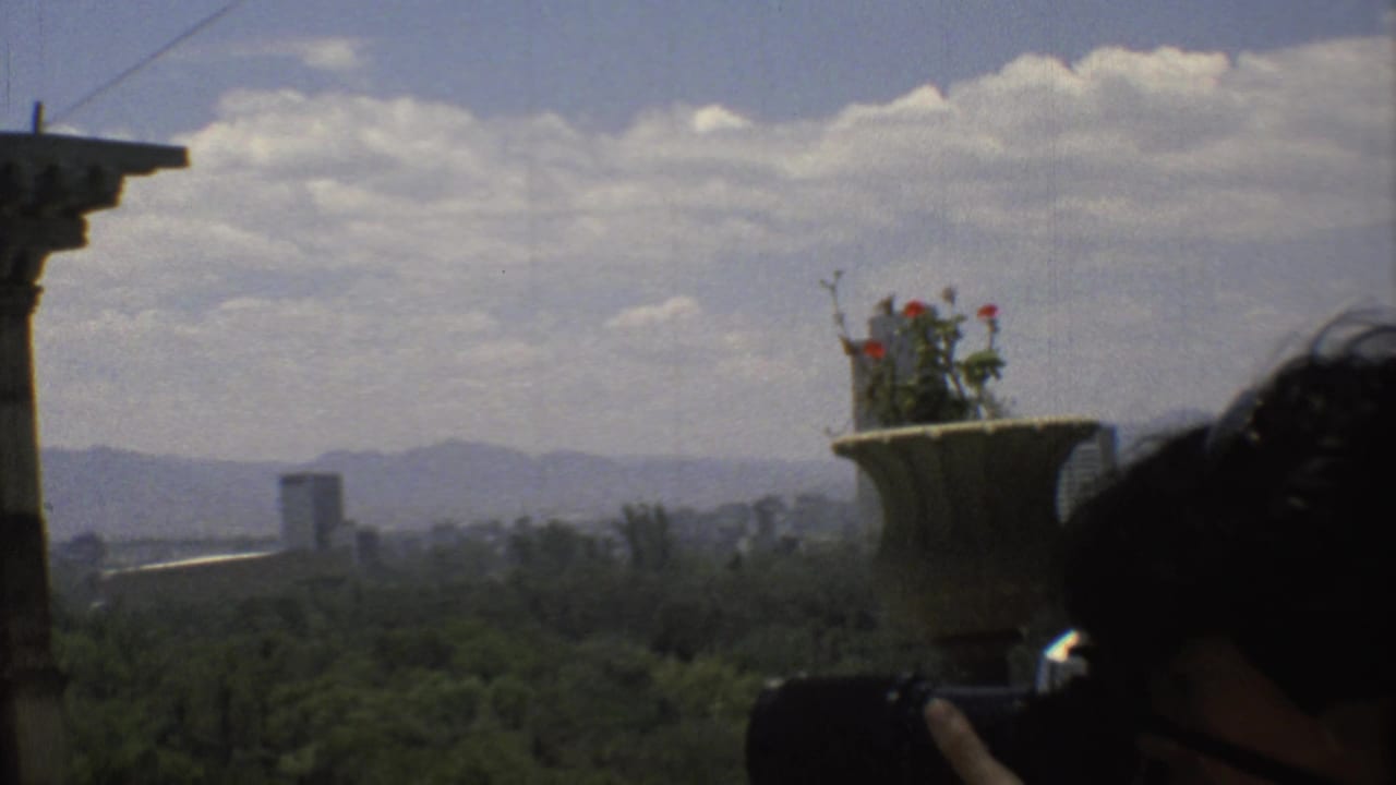 Handheld 8mm view over Mexico City with a stone porch edge, flower urn, distant buildings, mountains, and a partially visible person below frame.