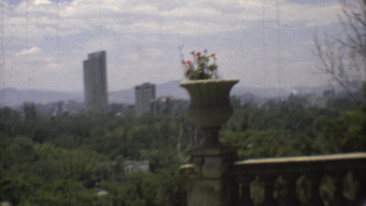 1973 Mexico City veranda view with a stone flower urn, balustrade, wooded foreground, modern towers, and hazy mountains.