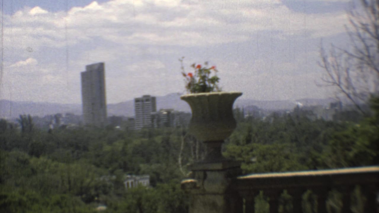1973 Mexico City home-movie view from a terrace, showing distant high-rises beyond greenery and mountains, framed by a stone urn.