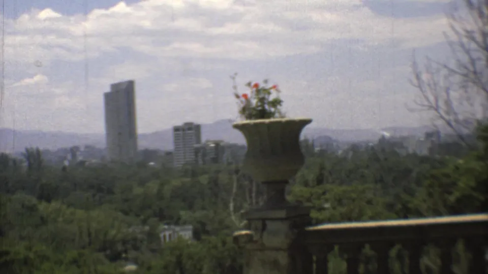 1973 Mexico City home-movie view from a terrace, showing distant high-rises beyond greenery and mountains, framed by a stone urn.