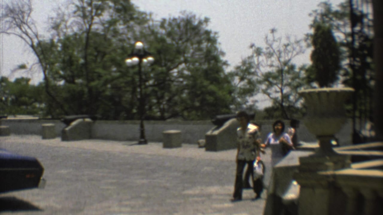 1973 Mexico City plaza scene with visitors walking near decorative urns, lampposts, cannons, trees, and a low stone wall.