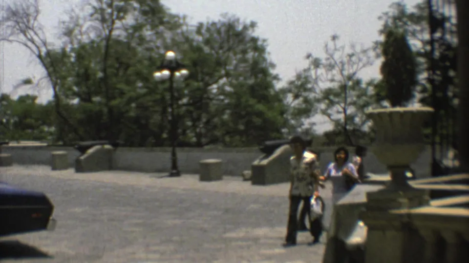 1973 Mexico City plaza scene with visitors walking near decorative urns, lampposts, cannons, trees, and a low stone wall.