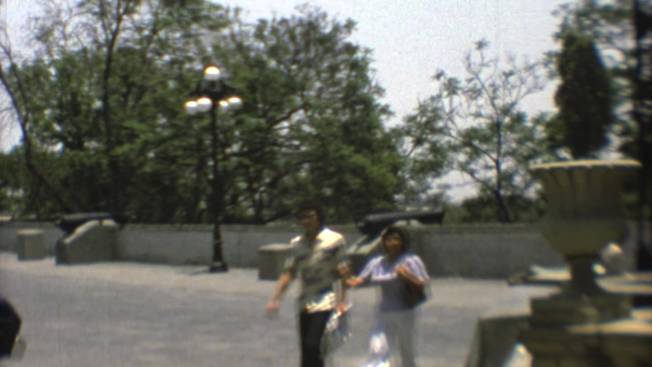 Mexico City home-movie frame from 1973 showing two adult visitors walking across a paved plaza with cannons and lampposts nearby.