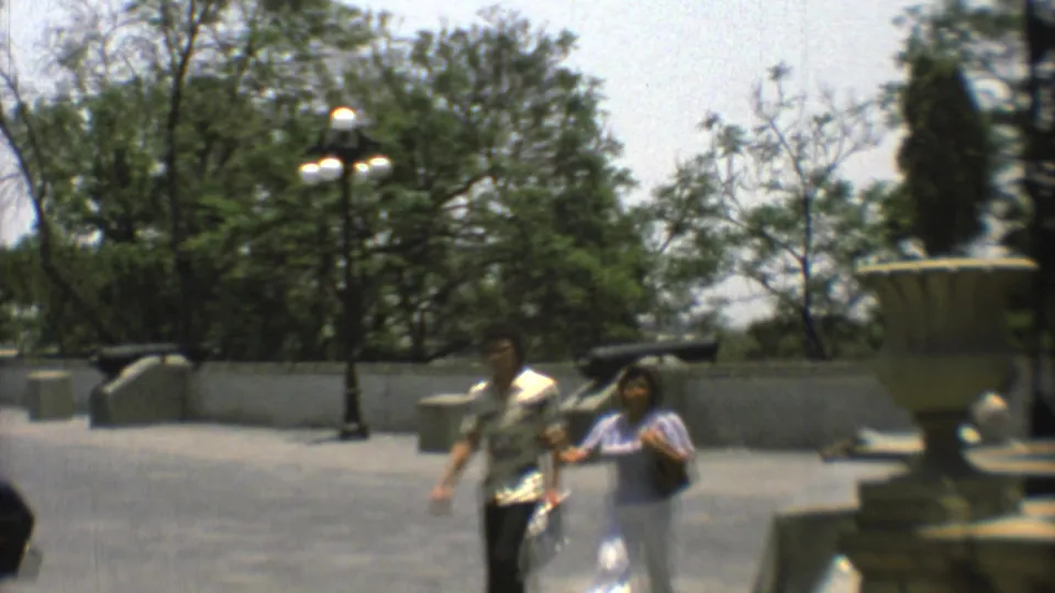 Mexico City home-movie frame from 1973 showing two adult visitors walking across a paved plaza with cannons and lampposts nearby.