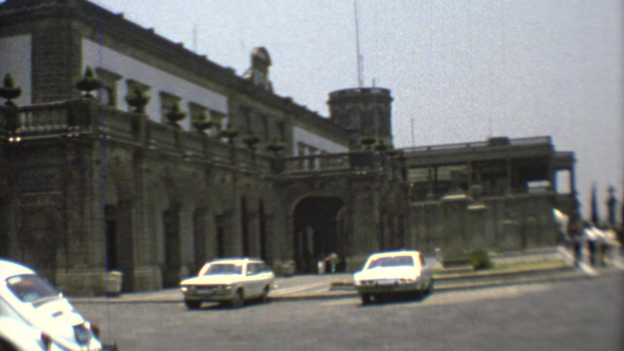 Historic stone building with arched entrance and cars passing on a sunlit street.