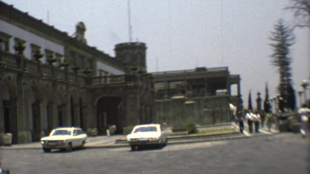Large classical building, two cars, and pedestrians near a gated walkway.