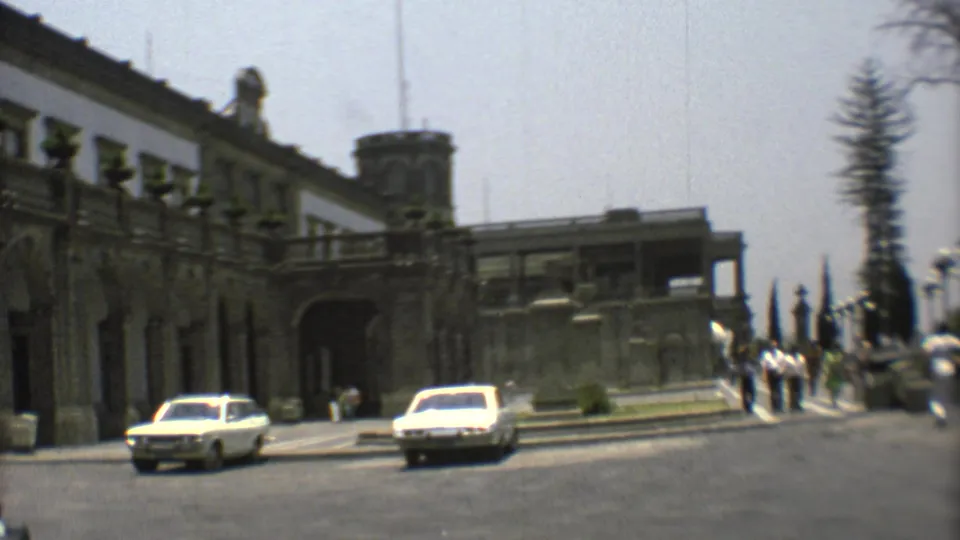 Historic Mexico City building exterior with a tower, arched entry, pedestrians, and 1970s cars moving through the foreground.