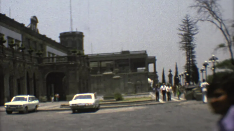 1973 Mexico City exterior view with a historic building, light-colored cars, distant pedestrians, and a blurred passerby close to camera.