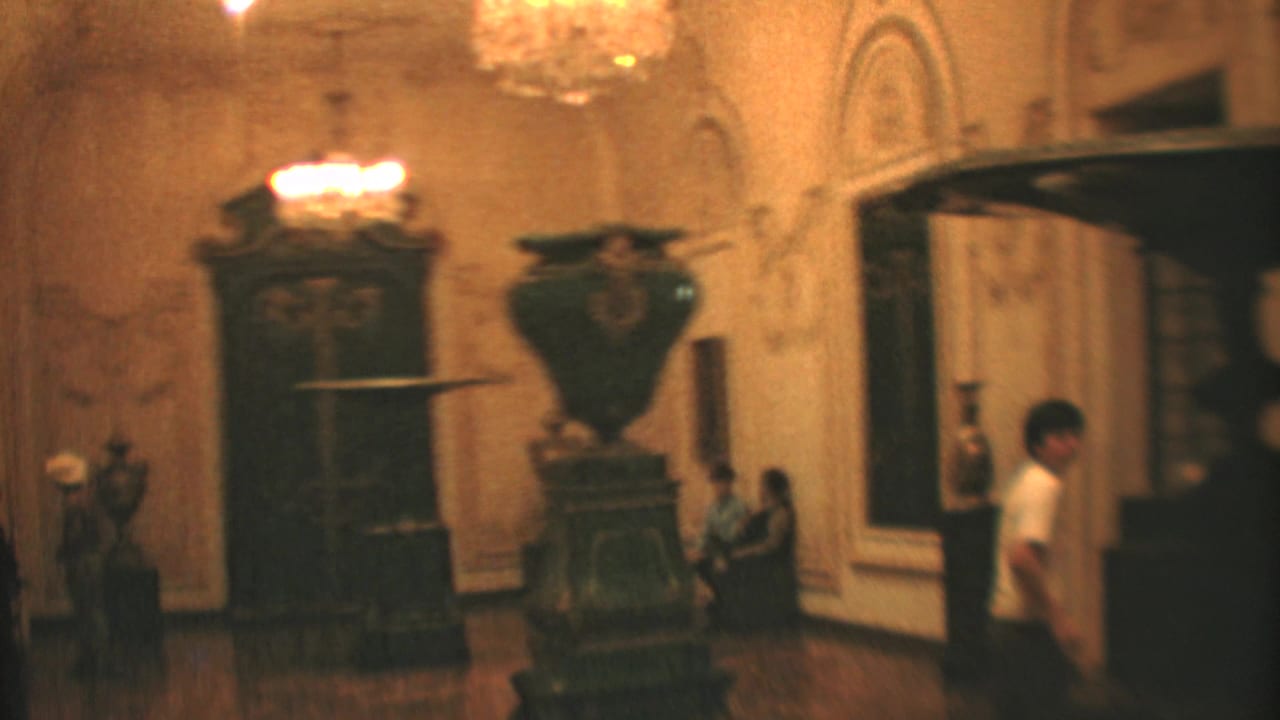 1973 Mexico City home-movie frame of an ornate interior where visitors sit and walk near display objects and a broad doorway.