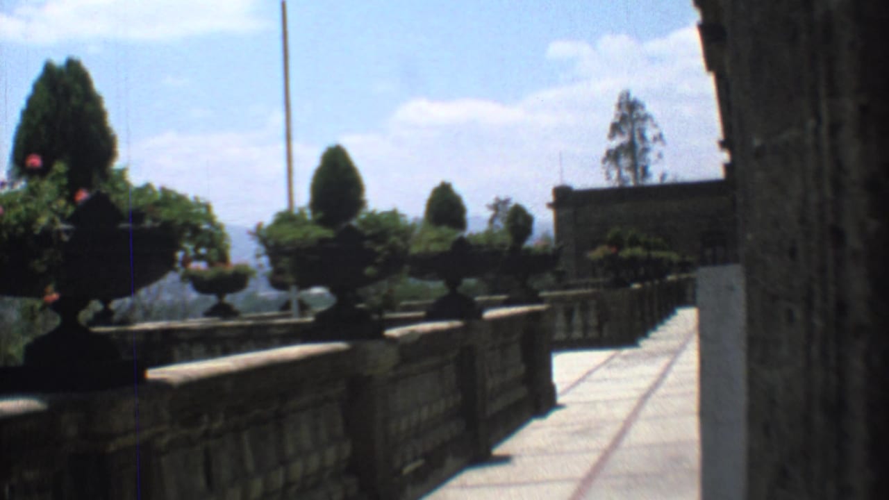 1973 Mexico City 8mm frame of a sunlit stone balcony with large planted urns, topiary, a flagpole, and a distant city view.