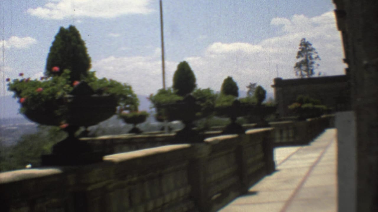 1973 Mexico City home-movie frame showing a paved balcony walkway with stone railings, formal urn planters, sculpted greenery, and sky beyond.