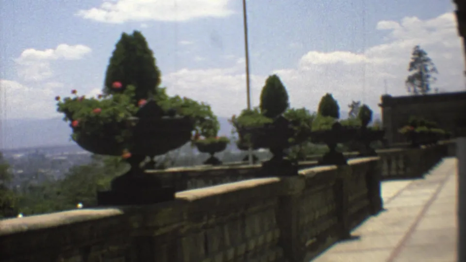1973 Mexico City 8mm frame centered on a large flowering urn beside a stone terrace, with topiary, flagpole, clouds, and city haze behind.