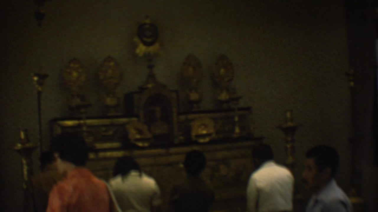 1973 Mexico City 8mm frame inside a dim room where visitors face an ornate golden altar or devotional display.