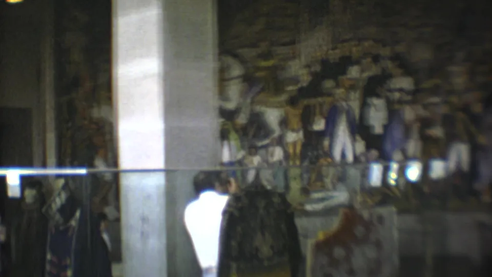 Visitors in a Mexico City museum stand near a glass display while a historical mural appears beyond a column.