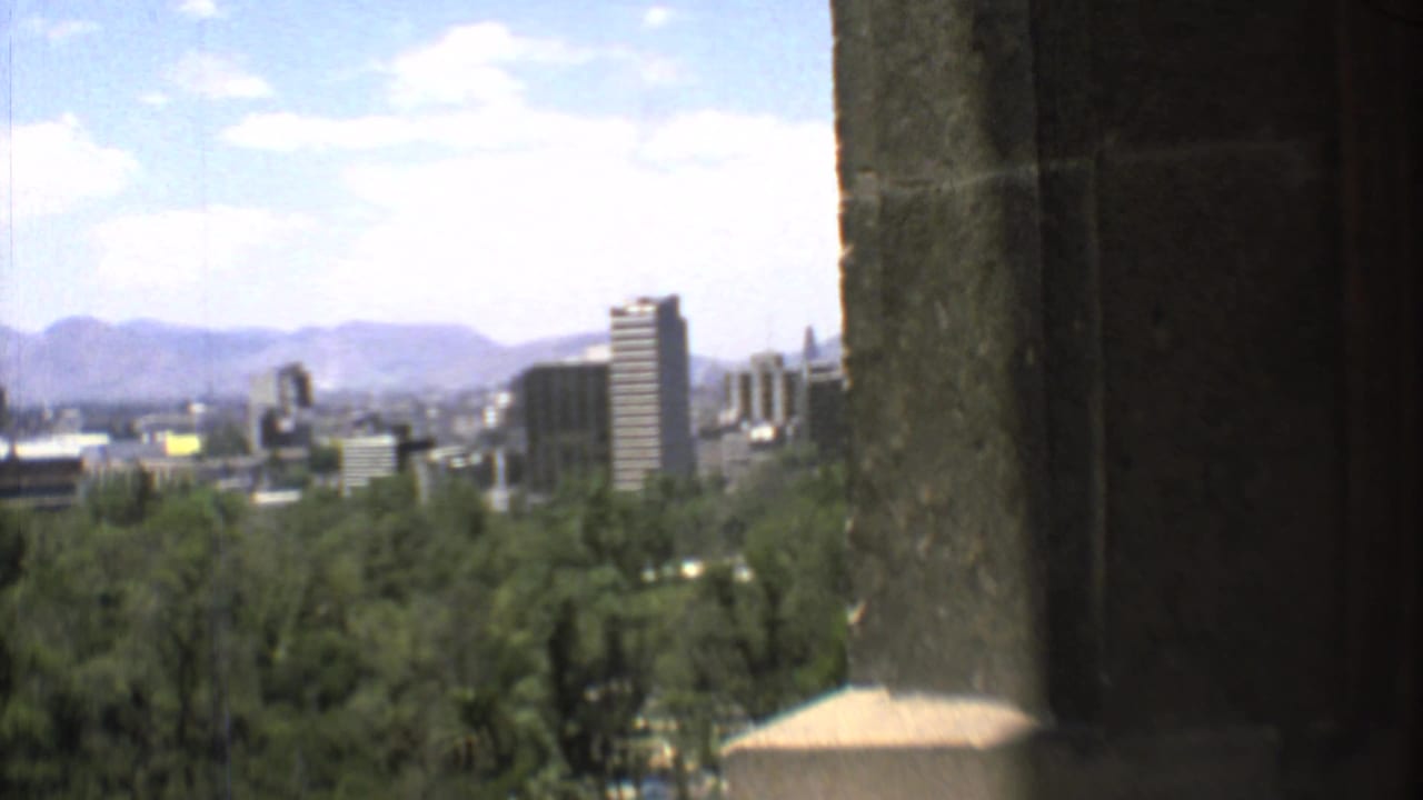 A high vantage view over Mexico City shows trees, mid-century buildings, distant mountains, and a partly cloudy sky.