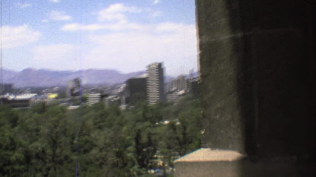 1973 8mm view over Mexico City with green treetops, mid-rise buildings, hazy mountains, blue sky, and a dark pillar at right.