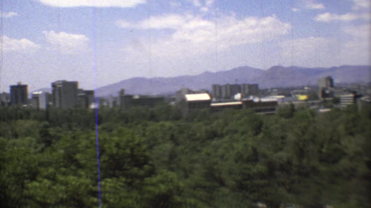 1973 8mm view of Mexico City skyline beyond dense green trees, with hazy mountains and scattered clouds in the distance.