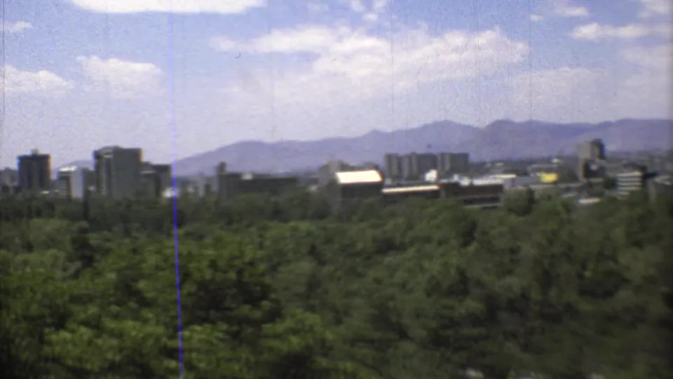 1973 8mm view of Mexico City skyline beyond dense green trees, with hazy mountains and scattered clouds in the distance.