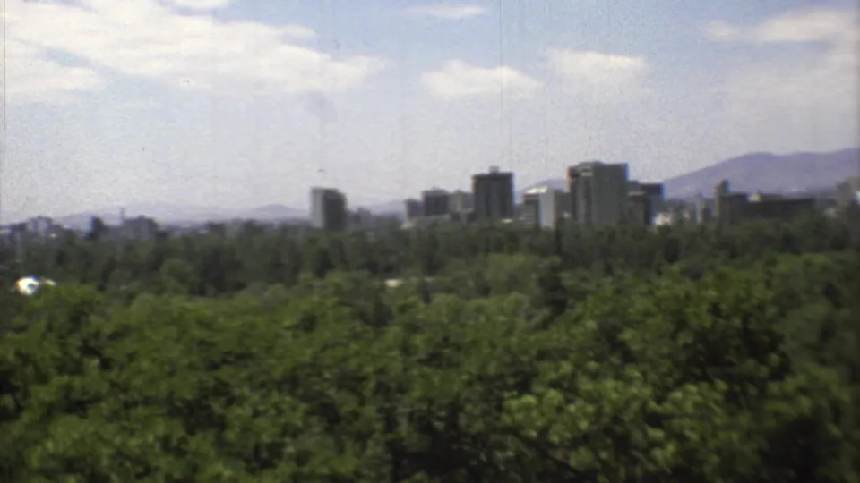 Wide 1973 home-movie view toward Mexico City's buildings, framed by green trees and a hazy mountain horizon.