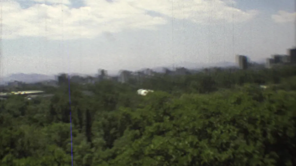 1973 Mexico City home-movie view showing trees in the foreground, distant towers, hazy mountains, and a pale clouded sky.