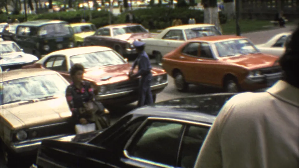 Mexico City street scene with 1970s sedans, parked cars, pedestrians, and trees captured in handheld 8mm footage.