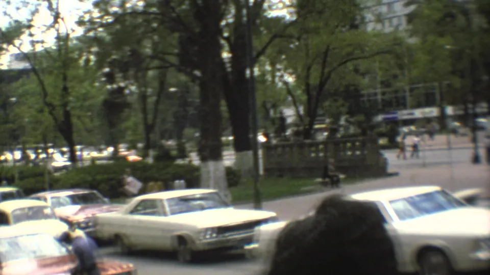 Mexico City street scene with a white sedan, parked cars, distant pedestrians, trees, and a paved public walkway.