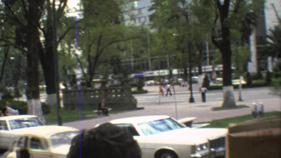 1973 Mexico City view across parked cars toward a sidewalk and small plaza, with pedestrians moving under trees.