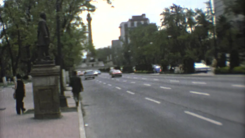 Mexico City avenue in 1973 with a sidewalk statue, pedestrians, multi-lane road, trees, and cars in the distance.