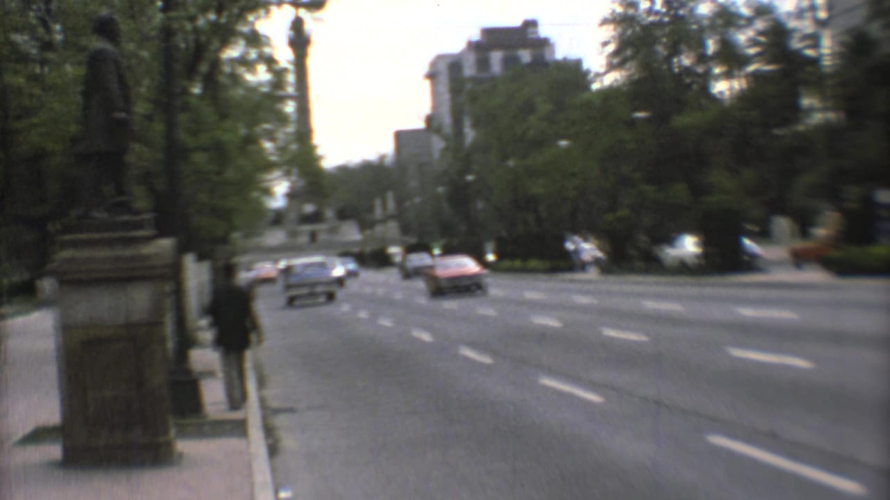 Tree-lined city avenue with cars passing beside a stone monument base and sidewalk.