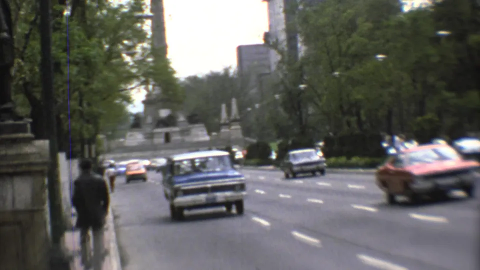 A visitor-shot 1973 Mexico City street view shows cars approaching near a sidewalk, trees, and a stone monument base.