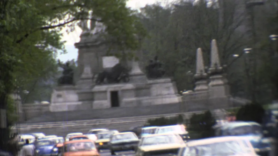 1973 Mexico City footage frames a large stone monument with lion sculptures, obelisks, trees, and passing cars.