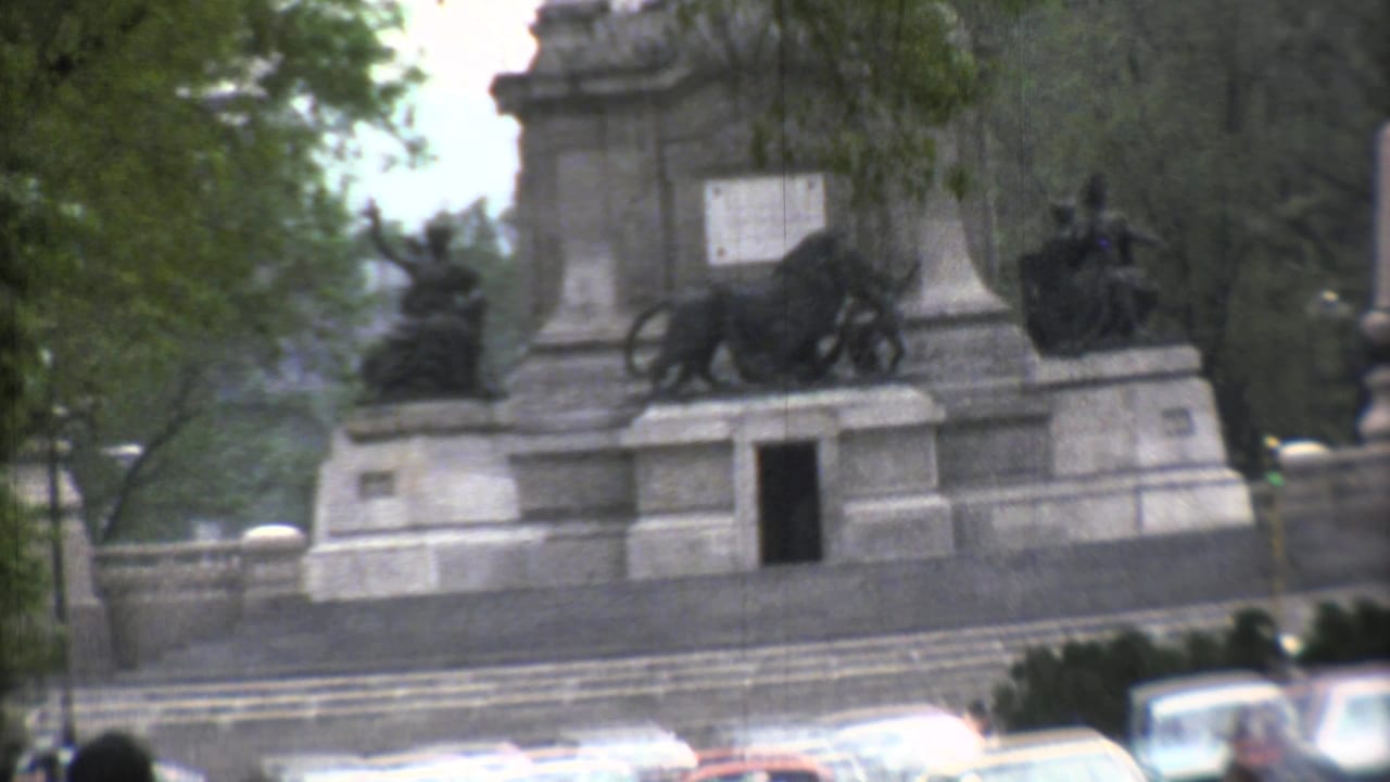 Large stone monument behind trees with classic cars moving along a city street.