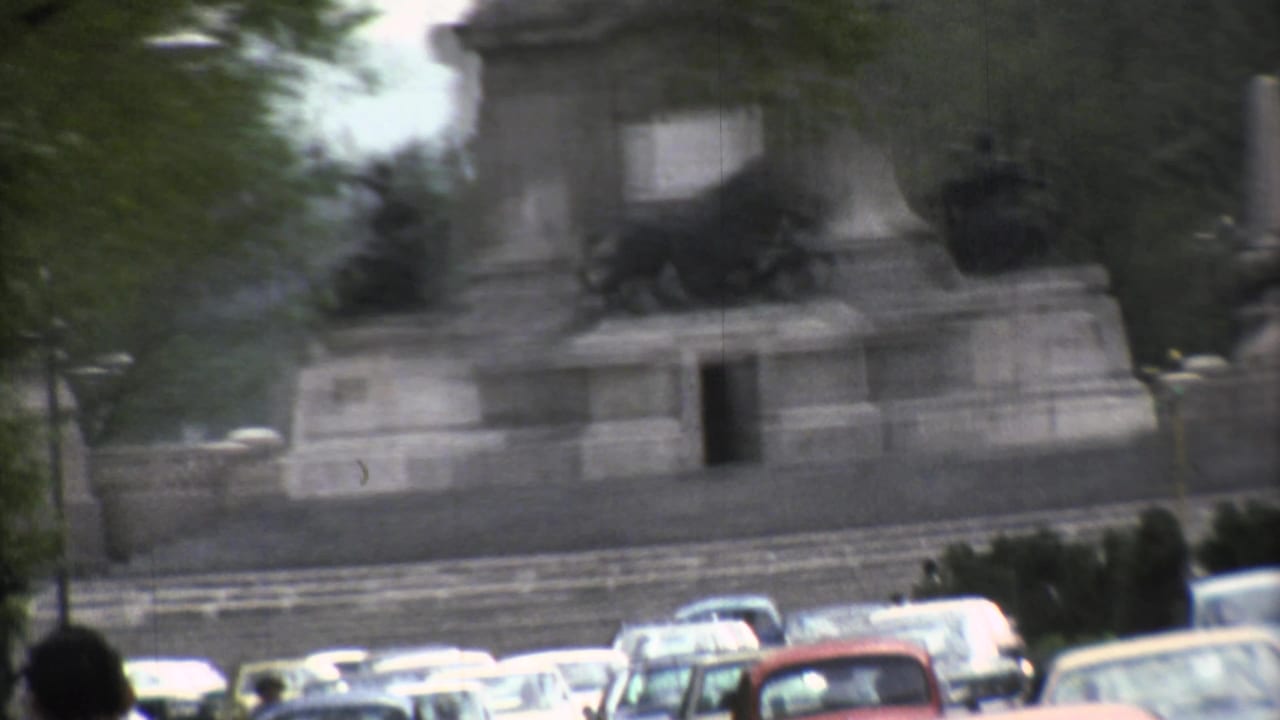 Soft 8mm frame of a Mexico City monument and tree-lined avenue with 1970s traffic passing below.