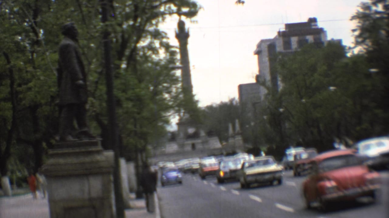A roadside statue frames a 1973 Mexico City traffic scene with trees, buildings, and classic cars beyond.