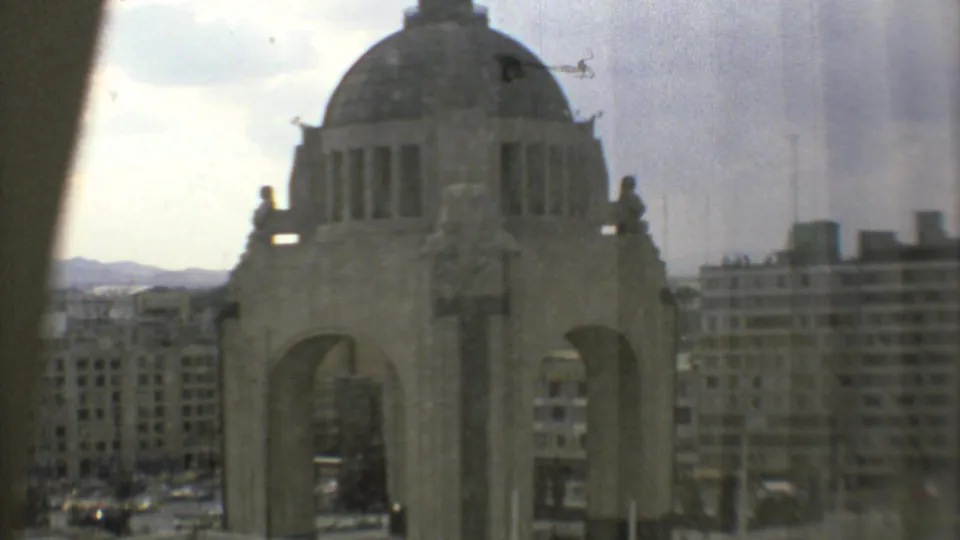 1973 Mexico City home-movie frame showing the Monumento a la Revolución from an elevated window, with apartment blocks and hazy sky behind it.