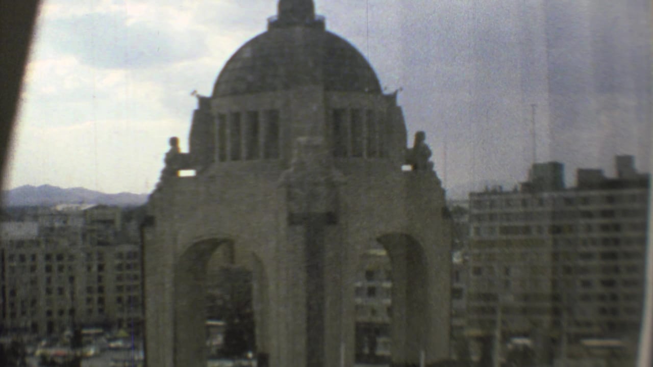 1973 Mexico City home-movie frame with the Monumento a la Revolución filling the foreground and modern buildings receding behind it.