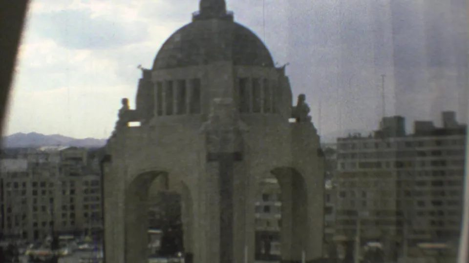 1973 Mexico City home-movie frame with the Monumento a la Revolución filling the foreground and modern buildings receding behind it.