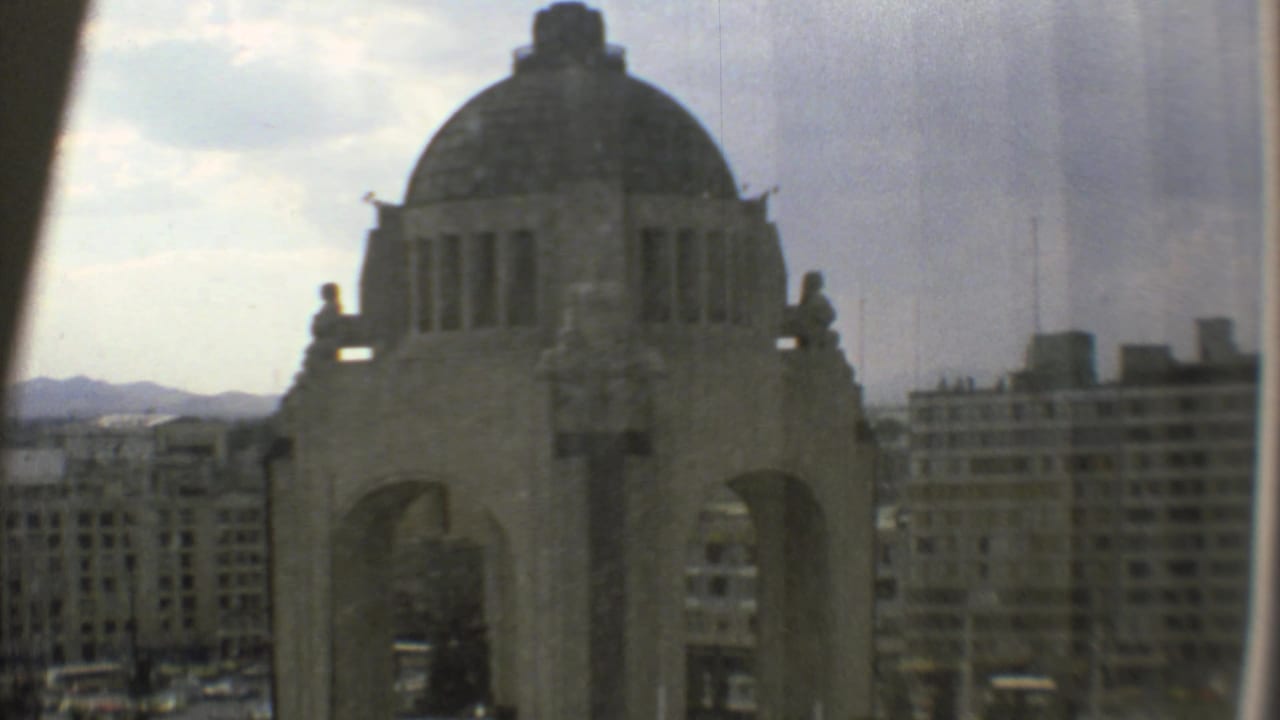 1973 Mexico City frame showing the Monumento a la Revolución through a window, with muted color, soft focus, and urban buildings beyond.