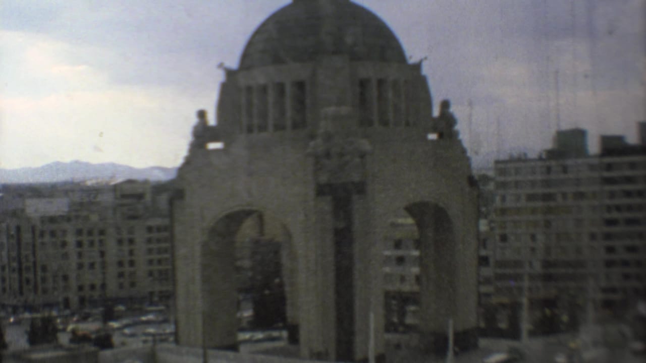 Domed stone monument with arches seen above city buildings and distant mountains.