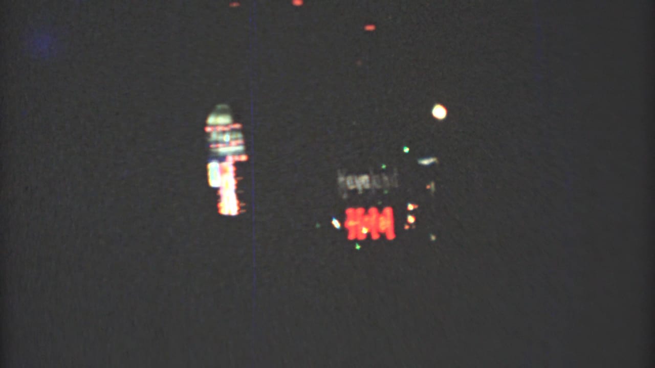 Dark nighttime street view with distant neon signs and small colored lights.