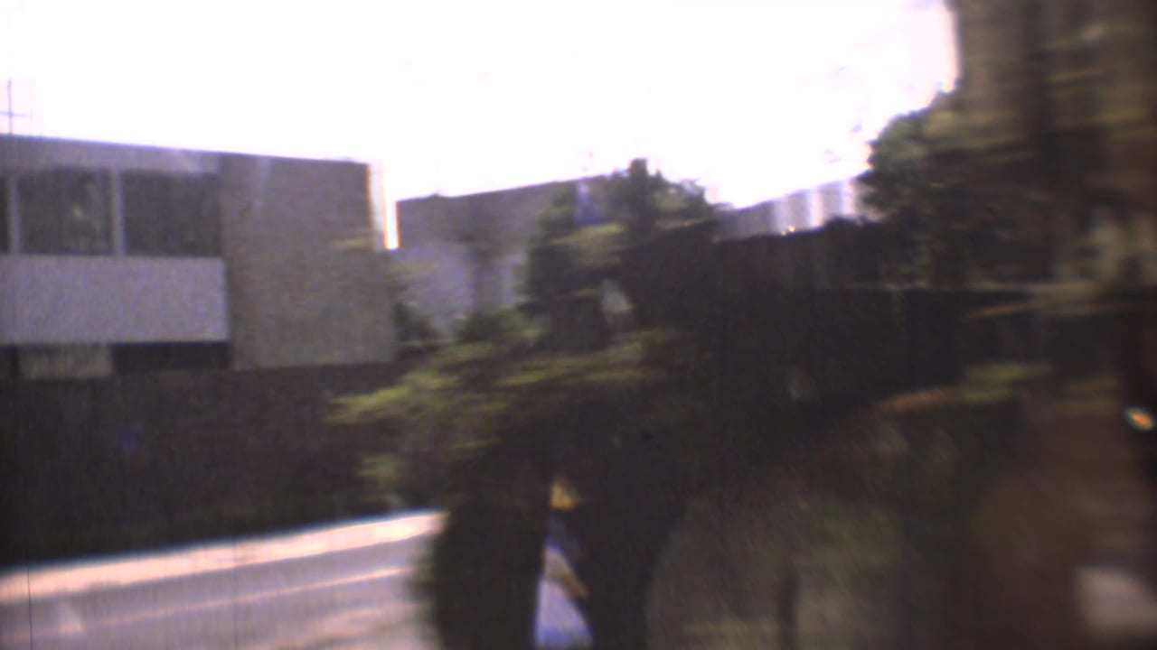 1973 Mexico City home-movie frame from inside a moving car, with a blurred street, building fronts, windshield edge, and roadside greenery.