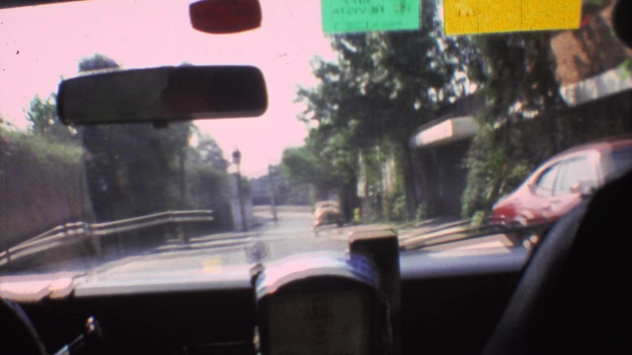 View through a car windshield on a sunny Mexico City street in 1973, with a rearview mirror, dashboard, trees, and a car ahead.