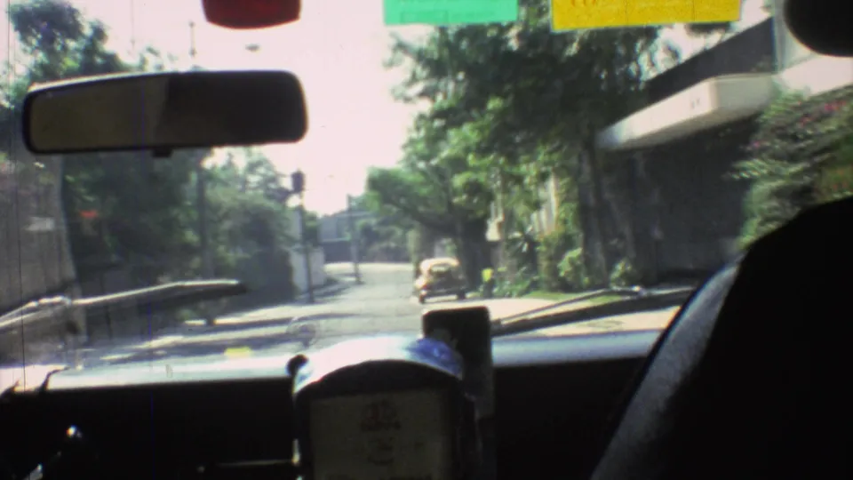 Inside-car view in Mexico City, 1973, looking past the rearview mirror and dashboard toward trees, buildings, and a car on the roadside.
