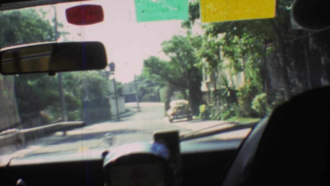 1973 Mexico City driving frame through the windshield, showing a leafy street, dashboard foreground, rearview mirror, and small car ahead.