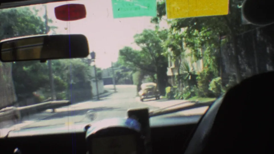1973 Mexico City driving frame through the windshield, showing a leafy street, dashboard foreground, rearview mirror, and small car ahead.