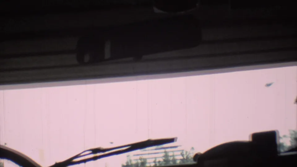 1973 Mexico City frame from inside a vehicle, with windshield wiper, dashboard shadow, pale wall, and low greenery visible outside.